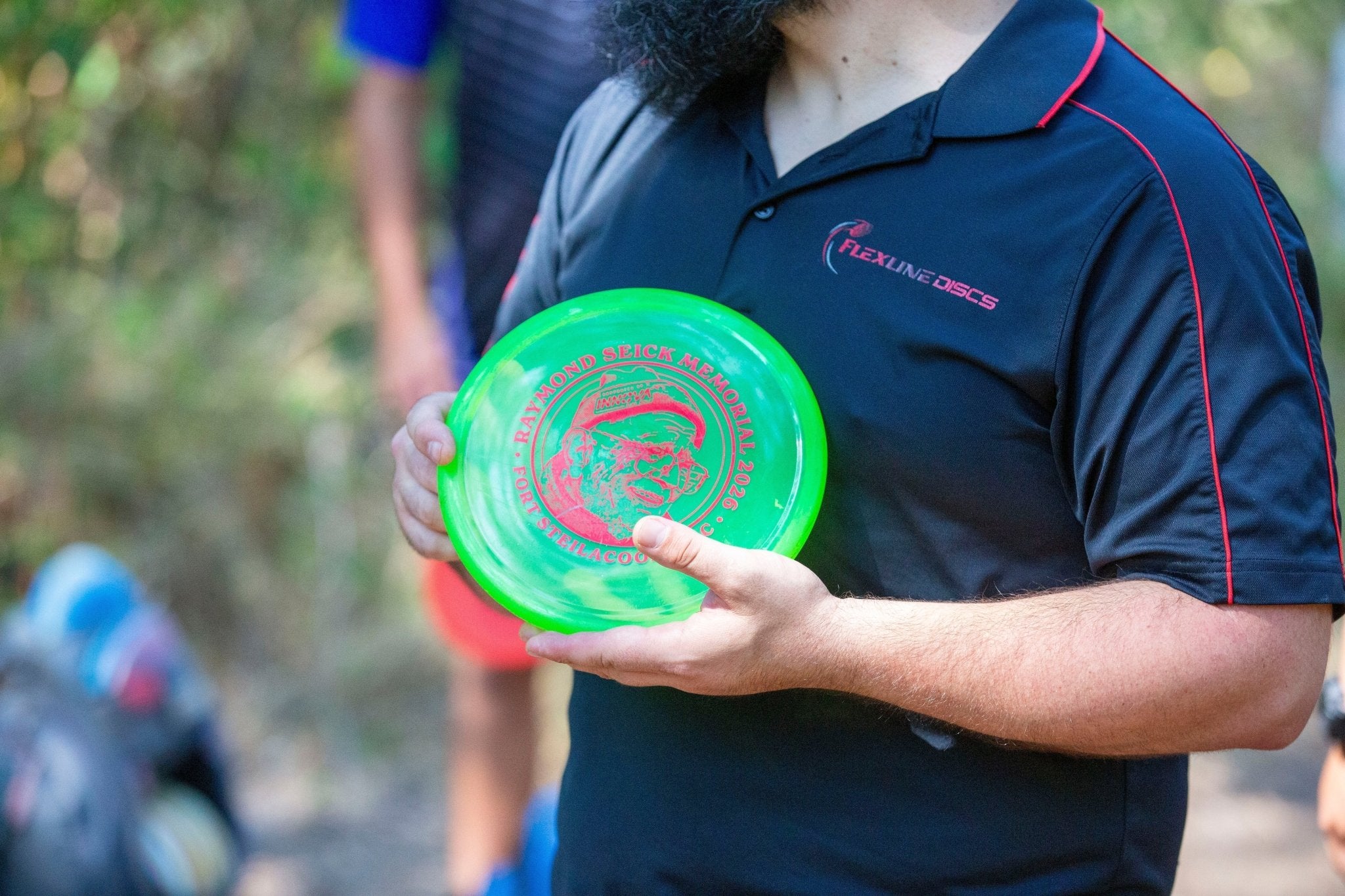 A man wearing a navy blue polo with the Flexline Discs logo on it. He is holding a green and red disc.
