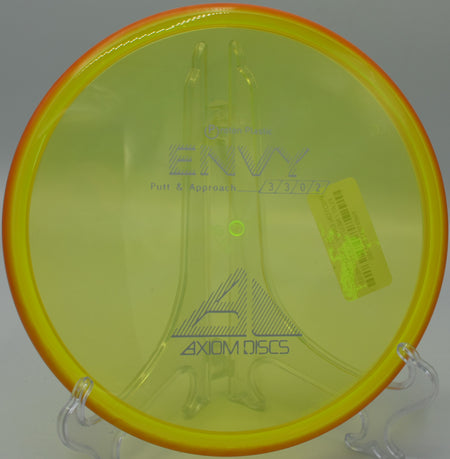 The Proton Envy displayed on a wooden bench, with sunlight reflecting off its smooth surface, taken at Maple Hill Disc Golf Course in Leicester, Massachusetts.