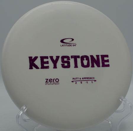 Zero Hard Keystone putter resting beside the basket at Maple Hill Disc Golf Course in Leicester, Massachusetts, featuring a firm feel and glide-heavy flight.

