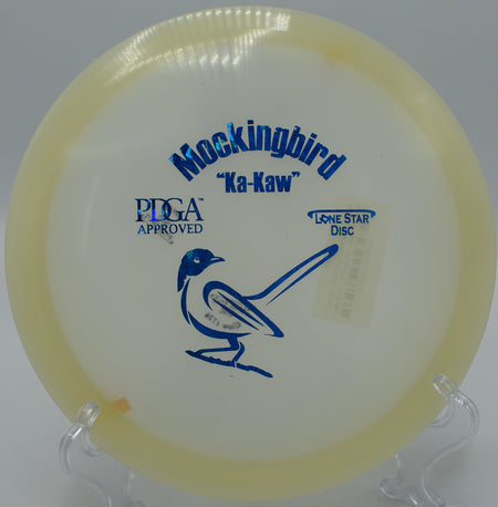 "Lone Star Glow Mockingbird resting near a basket at Rollin Ridge Disc Golf Course in Reedsville, Wisconsin, designed for consistent finishes."
